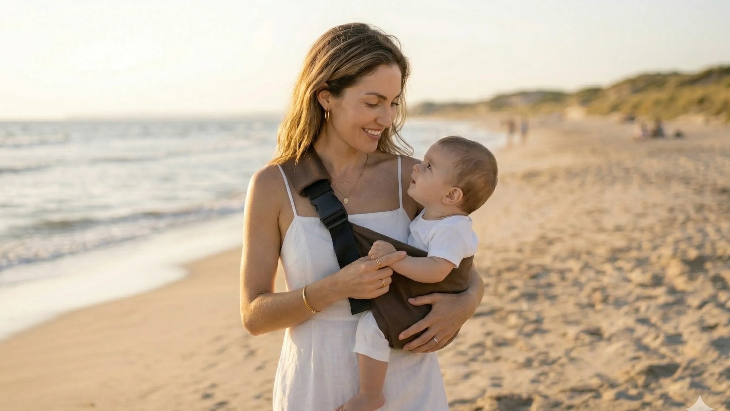 Madre sonriendo a su bebé en portabebés Bilby Moka marrón en la playa por la tarde