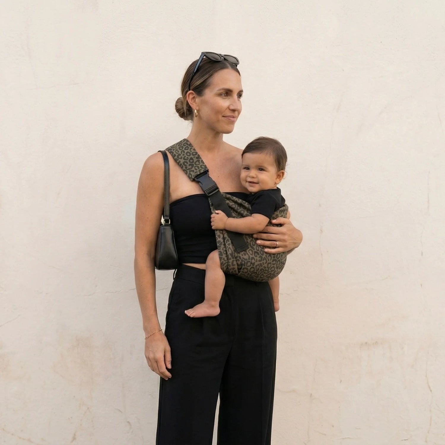 Madre con top negro elegante mirando al horizonte con bebé en portabebés Bilby Instinct leopardo — cuerpo entero fondo liso